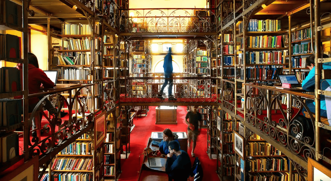 Cornell University library stacks with red carpet, two floors of bookshelves, and students studying and working on computers in the background.