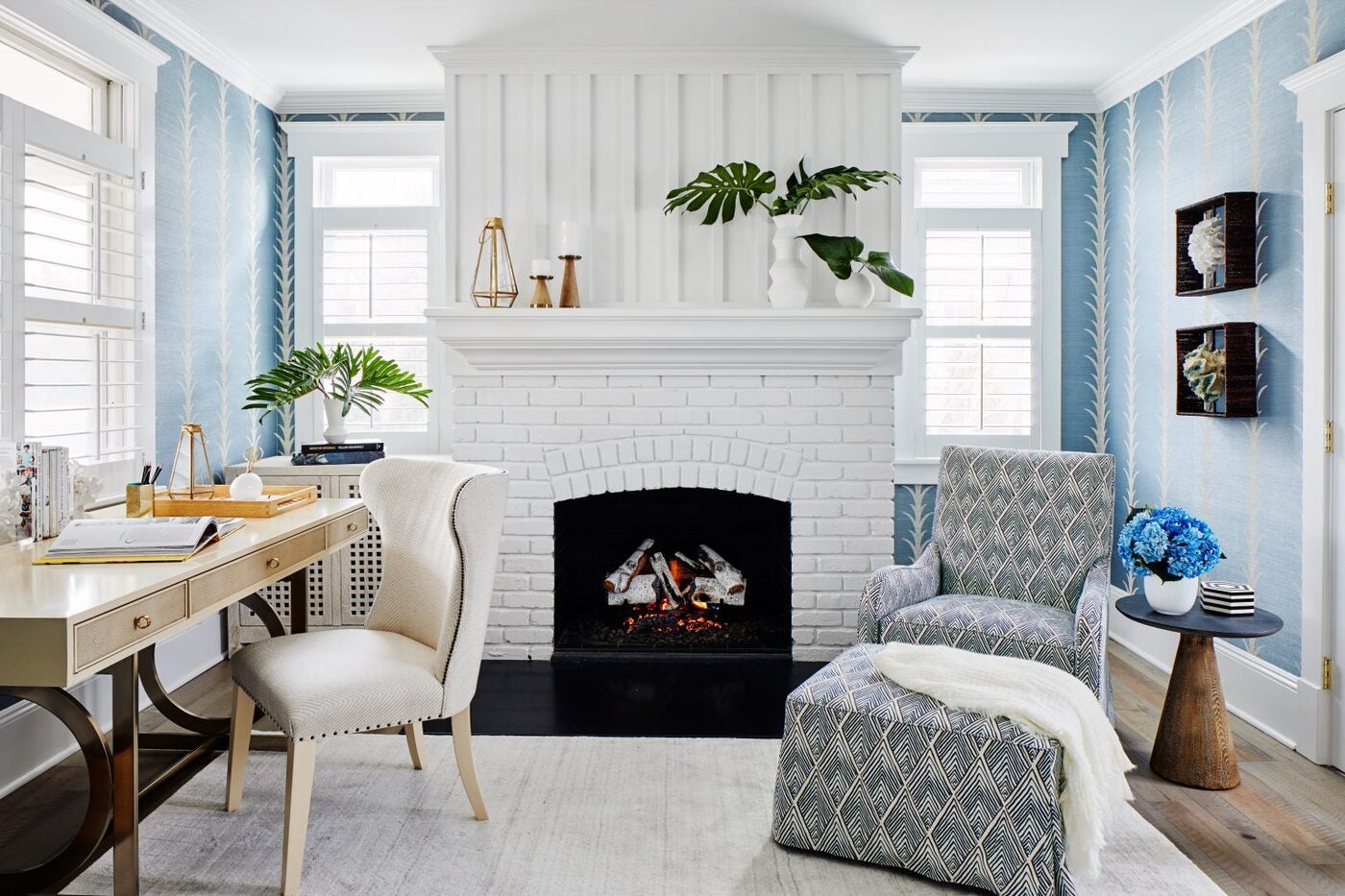 Brightly lit blue office, with a small wooden writing desk and tufted chair, white brick fireplace, and blue and white patterned armchair and ottoman.