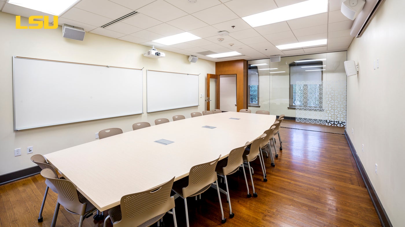 Large classroom at Louisiana State University with two whiteboards, a long table with rolling chairs, and glass doors.