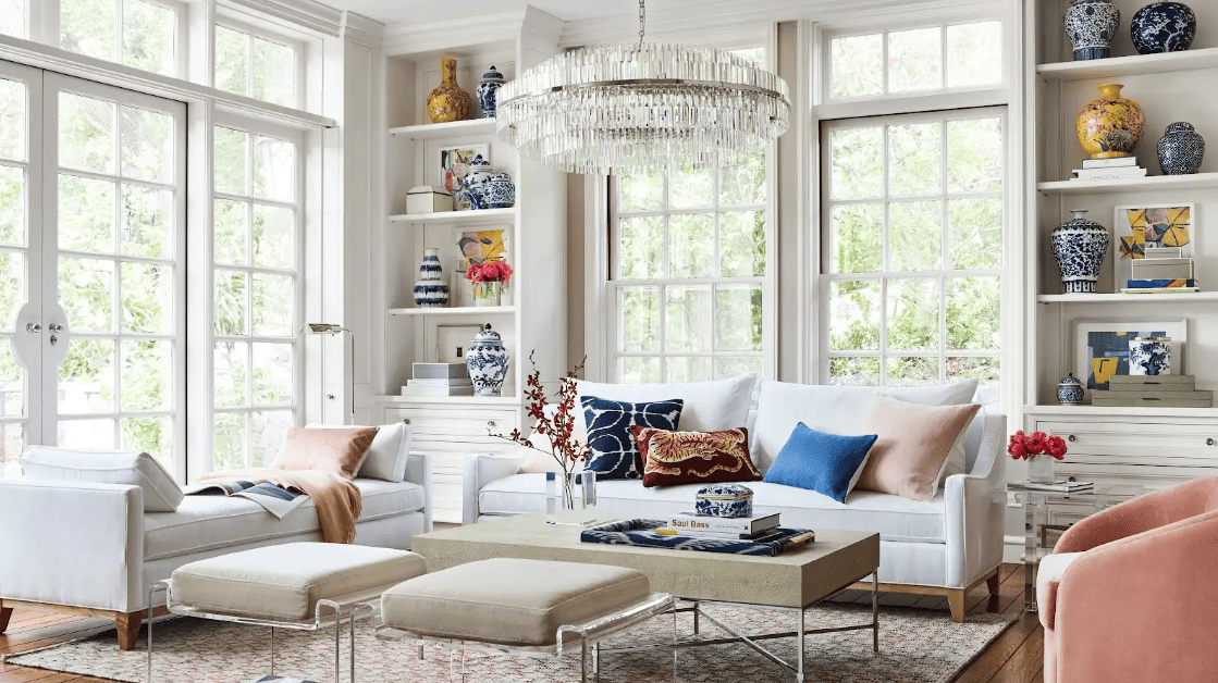 Sunlit living room with white floor-to-ceiling windows, built-in bookshelves, as well as white and pink accent furniture and pillows.