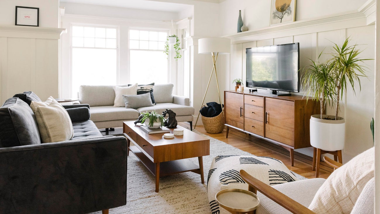 Mid-century modern living room with warm wood furniture, cream and grey suede couches, and a large sunlit window.