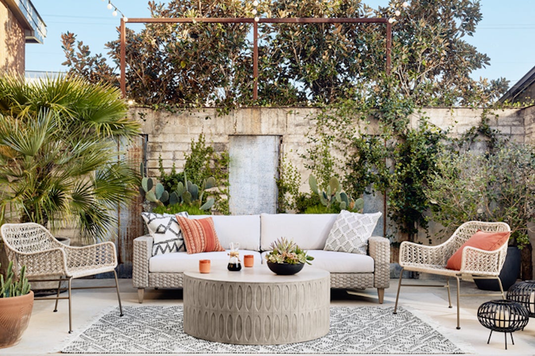 Outdoor patio with green shrubbery, a gray retaining wall, and wicker furniture with gray, coral, and cream pillows and accessories.