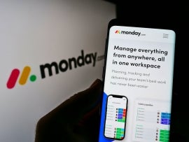 Person holding cellphone with webpage of company monday work management on screen in front of monday.com logo.