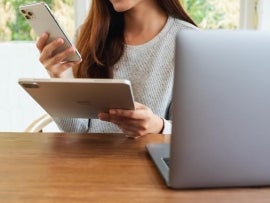 A person looking at an iPhone, holding a iPad and working on a Macbook.