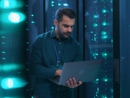 Caucasian IT professional admin using laptop computer doing data transfer operation with rack server cabinets in digital room of data center. Cyber security.
