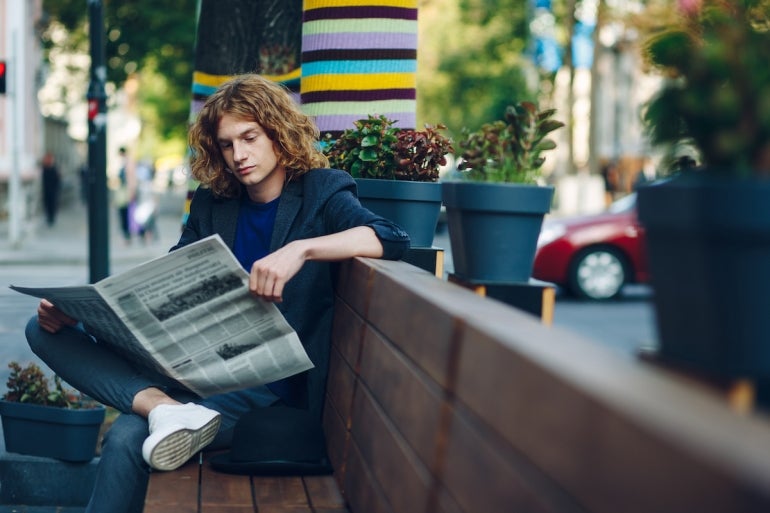 Red haired hipster man sitting on bench reading newspaper