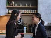 Two office workers chat in the break room with office supplies in the background.