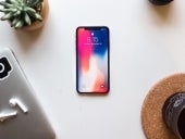 An iPhone sitting on a desk with a plant, a computer, and a cup.