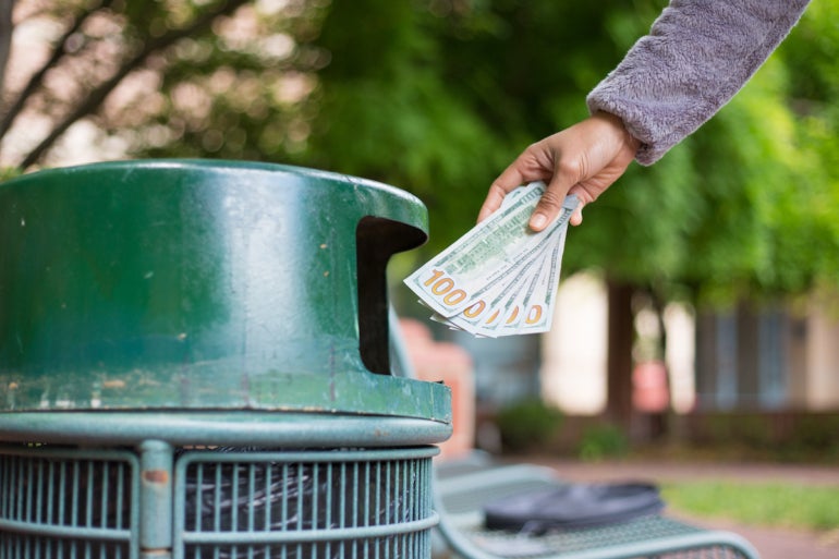 Closeup cropped portrait of someone hand tossing cash dollar bills money, hundred dollar bills in trash can, isolated outdoors green trees background. Bad financial investment decisions concept
