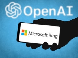 Shadowy hand holding a smartphone with Microsoft Bing on it over a blue background with the OpenAI logo.