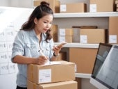 Woman making a note on top of a stack of boxes while looking at her phone