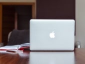 An Apple brand Macbook sits on a brown desk next to an open notebook.