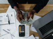 A Person Counting Us Dollars with a Macbook beside her desk.