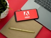 Adobe logo on the smartphone screen is placed on the Apple macbook keyboard on red desk background.