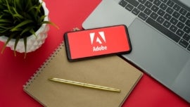 Adobe logo on the smartphone screen is placed on the Apple macbook keyboard on red desk background.