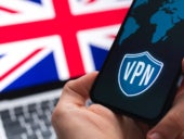 Human hand with mobile phone and VPN application with the flag of United Kingdom in the laptop screen as background.