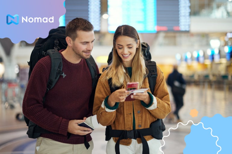 Two travelers wearing backpacks, standing in a transit center, look at a cell phone together.