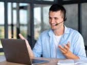 A happy man sitting in front of his laptop while wearing headphones and undergoing a virtual conference meeting.