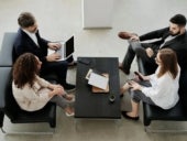 One person with a laptop, one person holding a cell phone, and two other business people sitting together in an office space.