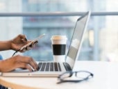 Person's hands coming into frame typing on a laptop and holding a mobile device with the other hand.