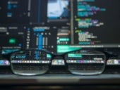 An out of focus computer screen with all sorts of applications open and a pair of glasses sitting on a table --- the screen you cans see through the glasses is in focus.