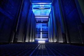 A modern data center server room with blue neon lighting, featuring tall, sleek server racks on both sides and a metal grid floor, representing web hosting infrastructure.