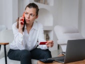 Worried businesswoman talks by phone while holding a credit card and sitting at desk with laptop.