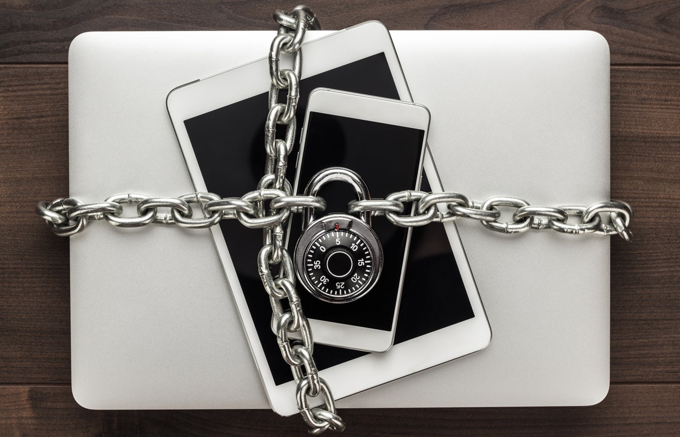 Computer, tablet, phone bound by metal chain and closed with combination lock on wooden table.