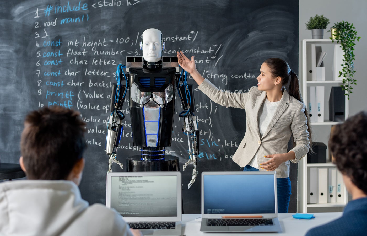 Clever girl standing by blackboard and pointing at robot while making presentation of his characteristics to her classmates at seminar.