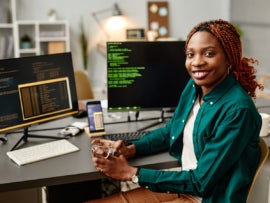 Portrait of female IT developer looking at camera and smiling against programming code on computer screen in office interior.