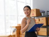 Woman holding clipboard smiling near stacked boxes in office