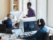 Young worker of cyber security center sitting in front of computers with graphic data and using smartphone while his colleagues having discussion