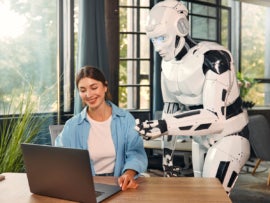 Robot and woman working on laptop in office.