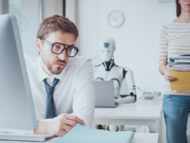 man wearing glasses and a tie sits at a desk, looking intently at a document in his hands, with a computer monitor in front of him. In the background, a humanoid robot is seated at another desk, seemingly working on a laptop, and a woman stands holding a stack of folders and papers. The scene suggests a modern office environment where human and artificial intelligence coexist and potentially interact in the workplace.