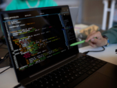 Focus on laptop screen with coded data and pen held by hand of young man.