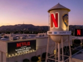 A digitally edited image of the Warner Bros. studio lot at sunset, featuring a large water tower that normally bears the Warner Bros. logo but is altered to display a large red Netflix logo alongside the WB shield. A nearby studio building has a large sign reading “NETFLIX ACQUIRES WARNER BROS. DISCOVERY.”