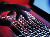 A hand illuminated in red light types on a backlit laptop keyboard, suggesting cyber activity, hacking, or intense computing