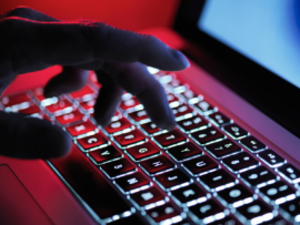 A hand illuminated in red light types on a backlit laptop keyboard, suggesting cyber activity, hacking, or intense computing
