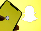 A person holding an iPhone displaying the Snapchat ghost logo on a bright yellow screen, with a larger Snapchat logo blurred in the background.