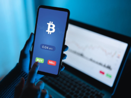A close-up of a person using a cryptocurrency trading app on a smartphone to buy or sell Bitcoin, with a financial market chart displayed on a laptop in the background.