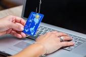 Credit card held by hand and punctured by a fishhook in front of a laptop.