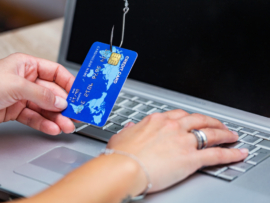 Credit card held by hand and punctured by a fishhook in front of a laptop.
