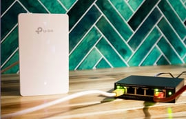 A white TP-Link Omada access point and a black TP-Link 5-port gigabit desktop PoE switch (TL-SG1005P) connected on a wooden surface with a green tile background.
