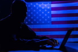 Hacker with laptop and USA flag in background.
