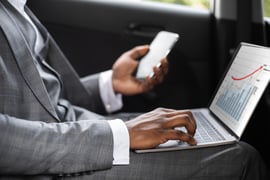 A man dressed professionally uses a smartphone and laptop inside his car.