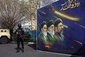 Armed security officer stands guard beside a large banner depicting Iranian leaders and a raised sword, with a military vehicle and trees in the background.