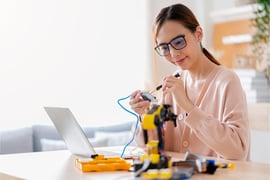 A young woman with glasses using a screwdriver to assemble a DIY robotic arm and circuit board at a desk with a laptop.