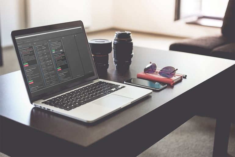 Laptop on a dark table showing code on the screen alongside camera lenses, a smartphone, and sunglasses.